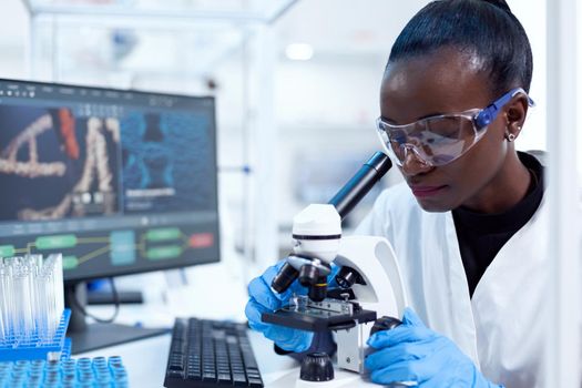 African pharmacology researcher analysing chemical substance using microscope for study.. Black healthcare scientist in biochemistry laboratory wearing sterile equipment.