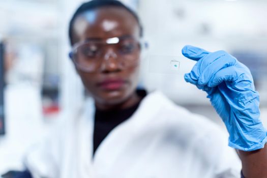 Sample glass slide in focus while chemist is looking at it during lab investigation. African healthcare scientist in biochemistry laboratory wearing sterile equipment.