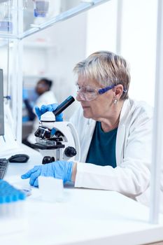 Senior scientist woman with protection glasses doing genetic investigation using microscope with african assistant in the background.. Elderly researcher carrying out scientific research in a sterile lab using a modern technology.