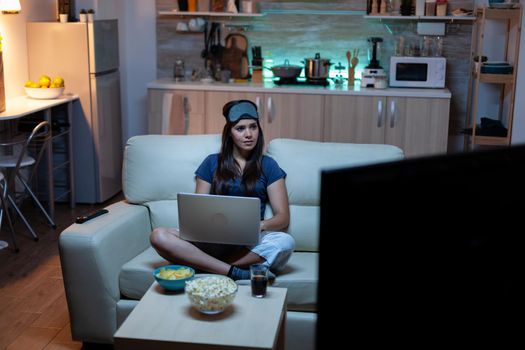 Young woman in pijamas working at night sitting on couch using laptop. Freelancer working in front of television reading writing searching browsing on notebook using internet technology