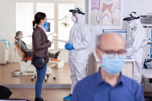 Stomatology doctor discussing with patient in reception dressed in ppe suit about health diagnosis. Medical dental speacilist during global pandemic with coronavirus.
