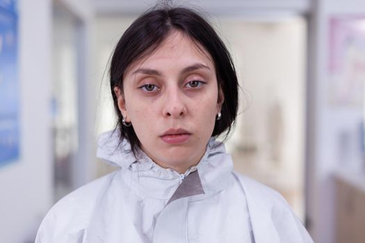 Close up of exhausted nurse in office looking on camera wearing ppe suit without face shield sitting on chair in waiting room clinic. Concept of new normal in coronavirus outbreak.