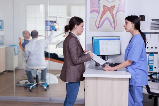 Woman in dentistiry clinic asking information to fill form for theeth treatment from receptionist. Doctor treating senior man in stomatology office. Examination radiograhy.