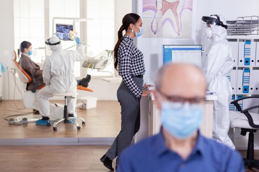 Stomatology patient with face mask discussing with dentist assitant wearing ppe suit face shield as safety precation during global pandemic with coronavirus. Dentistiry doctor consulting.