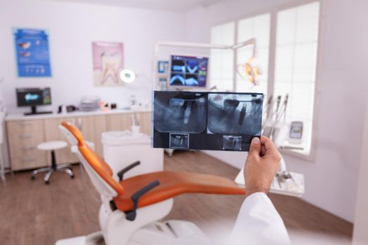 Specialist orthodontist looking at tooth dental radiograph working in stomatology medical hospital office room. In background empty orthodontic cabient preparing for teeth healthcare surgery