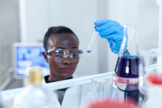 African scientist looking pensive at test tube with blue liquid. Black researcher in sterile laboratory conducting pharmacology experiment wearing coat.