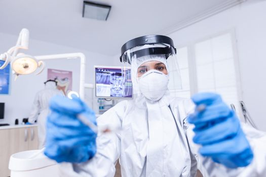 Patient pov of dentist examining patient gums using tools dressed up in covid suit in dental office. Stomatolog wearing safety gear against coronavirus during heatlhcare check of patient.