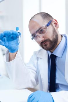 Male scientist with sterile gloves looing at test tube with genetic material. Researcher in biotechnology sterile lab holding analysis in tube wearing gloves and protection glasses.