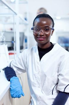 African scientist wearing protective glasses in medical laboratory looking at camera. Multiethnic team of researchers working in microbiology lab testing solution for medical purpose.