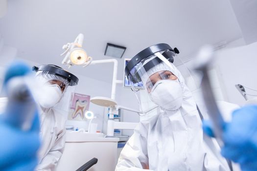 Patient pov of dental team leaning over to work in teeth cavity dressed in coverall. Stomatology team wearing safety equipment against coronavirus treating patient.