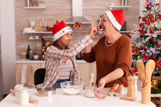 Happy family enjoying winter season putting flour on grandma nose while preparing homemade gingerbread dough in xmas decorated kitchen. Child cooking traditional cookies celebrating christmas holiday