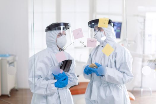 Dentist coworkers talking about teeth issues wearing pee suit as safety precauiton for covid-19. Medical team in stomatology office wearing coverall in dental office writing ideas on sticky notes.