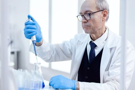 Elderly scientist doing pharmaceutical experiment using dropper pipette. Senior professional chemist using pippete with blue solution for microbiology tests.