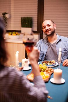 Guy celebrating relationship in kitchen with happy wife holding glass of red wine walking happy sitting at table dining room, enjoying the meal at home having romantic time at candle lights