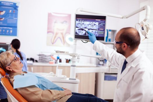 Stomatolog explaining diagnosis to senior woman during dental examination. Medical teeth care taker pointing at patient radiography on screen sitting on chair.