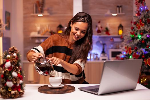 Festive adult using video call technology on laptop and pouring cup of tea preparing for winter season at home. Cheerful young woman talking to family on internet online in decorated room