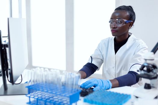 African biotechnology researcher works in bright modern laboratory using computer. Multi ethnic healthcare scientist in biochemistry facility wearing sterile equipment.