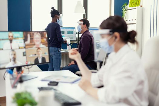 Man discussing with african coworker about a project at workplace wearing face mask against covid. Entrepreneur having videocall while colleagues working respecting social distance during global pandemic.