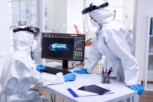 Dental team in ppe suit examining patient teeth x-ray. Medical specialist wearing protective gear against coronavirus during global outbreak looking at radiography in dental office.