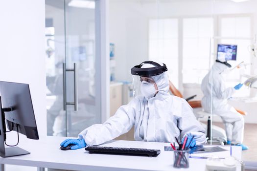 Medicine practitioner dressed in pee suit during covid 19 as safety precaution using computer. Medicine team wearing protection gear against coronavirus pandemic in dental reception as safety precaution.