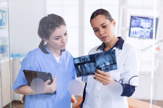 Orthodontist and nurse in stomatology office holding teeth x-ray wearing uniform. Stomatolog and her assistant in reception of dental clinic looking at teeth radiography.