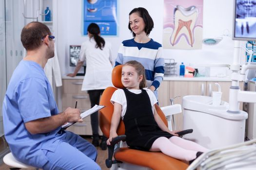 Stomatolog assistant discussing about tooth hygine of kid with parent in dental office. Child with her mother during teeth check up with stomatolog sitting on chair.