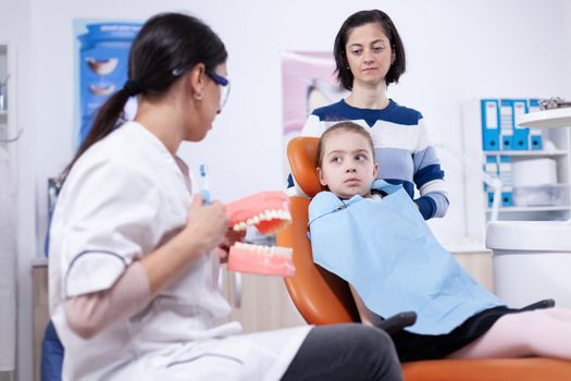 Stomatologist in the course of teeth brushing artificial jaw in the course of child check up. Little girl and mother listening stomatolog talking about tooth hygine in dentistiry clinic holding jaw model.