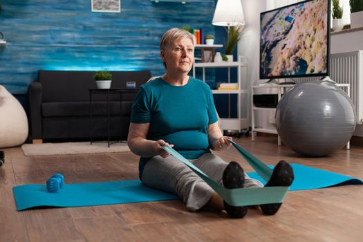 Retirement senior woman sitting on yoga mat stretching legs muscles using stretch elastic band training body resistance. Pensioner in sportswear slimming weight during muscle training in living room