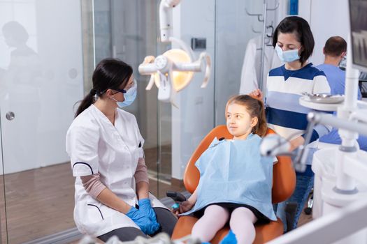 Dentist wearing face mask in the course kid examination with dental bib in dentistiry office. Dentistry specialist during child cavity consultation in stomatology office using modern technology.