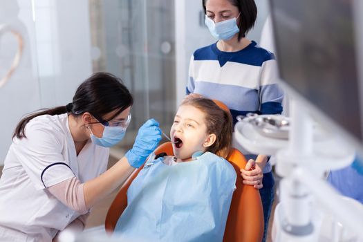 Dentist wearing gloves during cavity procedure on little girl sitting on dental chair. Dentistry specialist during child cavity consultation in stomatology office using modern technology.