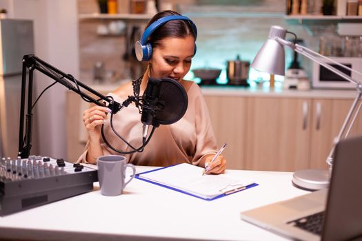 Presenter writing on clipboard and wearing headphones during podcast recording using professional microphone. Creative online show On-air production internet broadcast host streaming live content