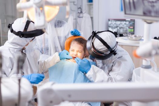 Little girl with eyes closed because of pain in the course of cavity treatment. Stomatology team wearing ppe suit during covid19 doing procedure on child teeth.