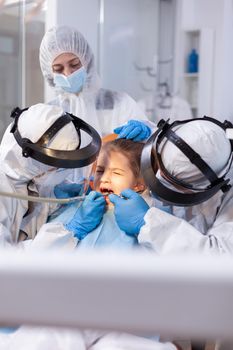 Little girl at dentist's appointment having a painfull tooth treatment dressed in protective suit against covronavirus. Stomatology team wearing ppe suit during covid19 doing procedure on child teeth.