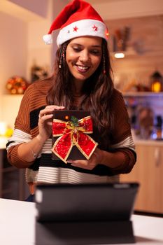 Young person using video call technology on tablet to give seasonal christmas present in festive decorated kitchen. Caucasian woman sharing winter joy with friends preparing for festivity