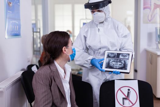 Stomatologist in protective suit pointing on digital x-ray of tooth explaining to patient treatment using tablet in covid-19 pandemic. Medical team wearing face shield, coverall, mask and gloves.