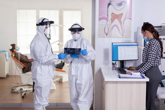 Stomatology team dressed up in ppe suit during global pandemic with coronavirus in dental reception holding patient x-ray, keeping social distancing. Receptionist with face mask as prevention.