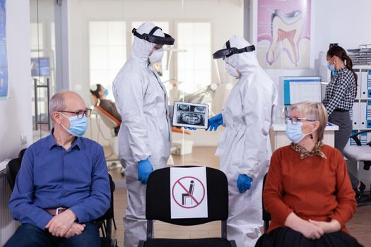 Man discussing with nurse in dental reception wearing protection suit against coronavirus, elderly patients waiting in reception keeping distance. Concept of new normal dentist visit in outbreak.