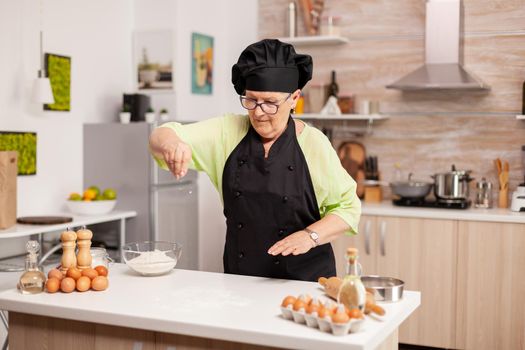 Retired chef preparing pizza in home kitchen wearing apron. Happy elderly chef with uniform sprinkling, sieving sifting raw ingredients by hand in home kitchen.