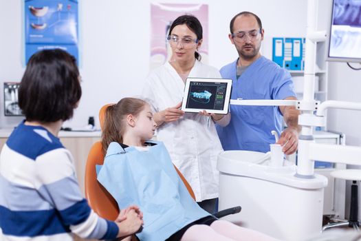 Dentist in dentistiry office showing tablet pc with jaw radiography explaining cavity treatment. Stomatologist explaining teeth diagnosis to mother of child in health clinic holding x-ray.