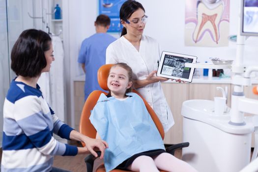 Dentist discussing diagnosis with child parent holding tablet pc with radiography. Stomatologist explaining teeth diagnosis to mother of child in health clinic holding x-ray.