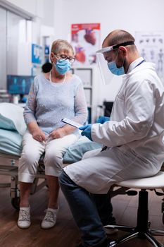 Doctor reading diagnosis of senior woman illness wearing face mask against covid pandemic as safety precaution in hospital. Healthcare medical physician consultation during COVID-19 global crisis.