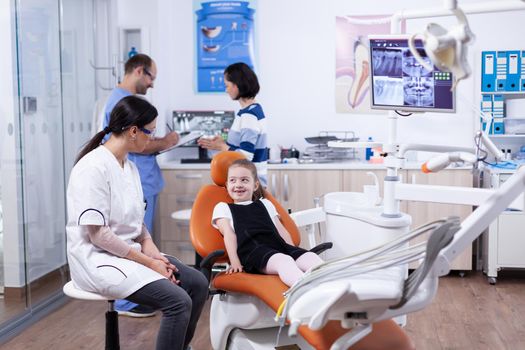 Little girl in stomatology office for teeth treatment looking at dentist. Child with her mother during teeth check up with stomatolog sitting on chair.