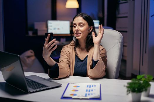 Businesswoman waving in the course of call with colleagues doing overtime. Woman working on finance during a video conference with coworkers at night hours in the office.