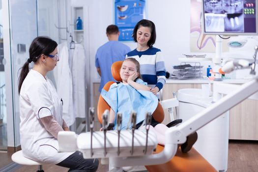 Little girl in pediatric dentist office touching face with painful expression because of tooth health issues. Child with her mother during teeth check up with stomatolog sitting on chair.