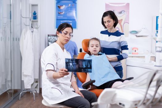 Little girl and parent listening dentist advice pointing at dental radiography. Stomatologist explaining teeth diagnosis to mother of child in health clinic holding x-ray.