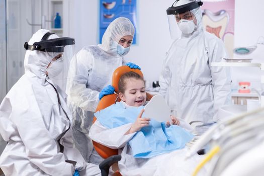 Little patient holds mirror in the course of oral hygine treatment.Child wearing ppe suit during teeth intervention at dental hospital.