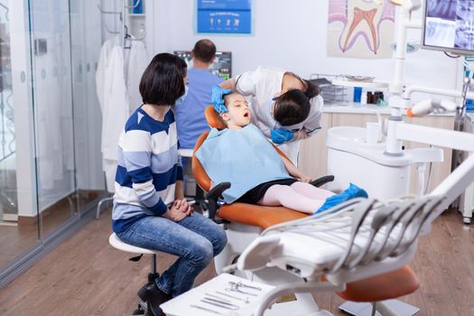 Little girl in the course of treatment for caries sitting on dental chair wearing bib. Dentistry specialist during child cavity consultation in stomatology office using modern technology.