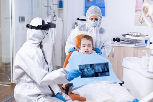 Little girl and mother wearing ppe suit looking at dental radiography in dentist office during covid19 pandemic. Stomatolog in protectie suit for coroanvirus as safety precaution holding child teeth x-ray during consultation.