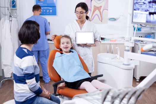 Dentist in clinic with chroma key on tablet pc display consulting kid with bad tooth. Stomatolog explaining teeth prevention to mother and child holding tablet pc with copy space available.