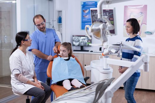 Kid patient in dentist office prepared for stomatology treatment while parent is talking with doctor. Child with her mother during teeth check up with stomatolog sitting on chair.
